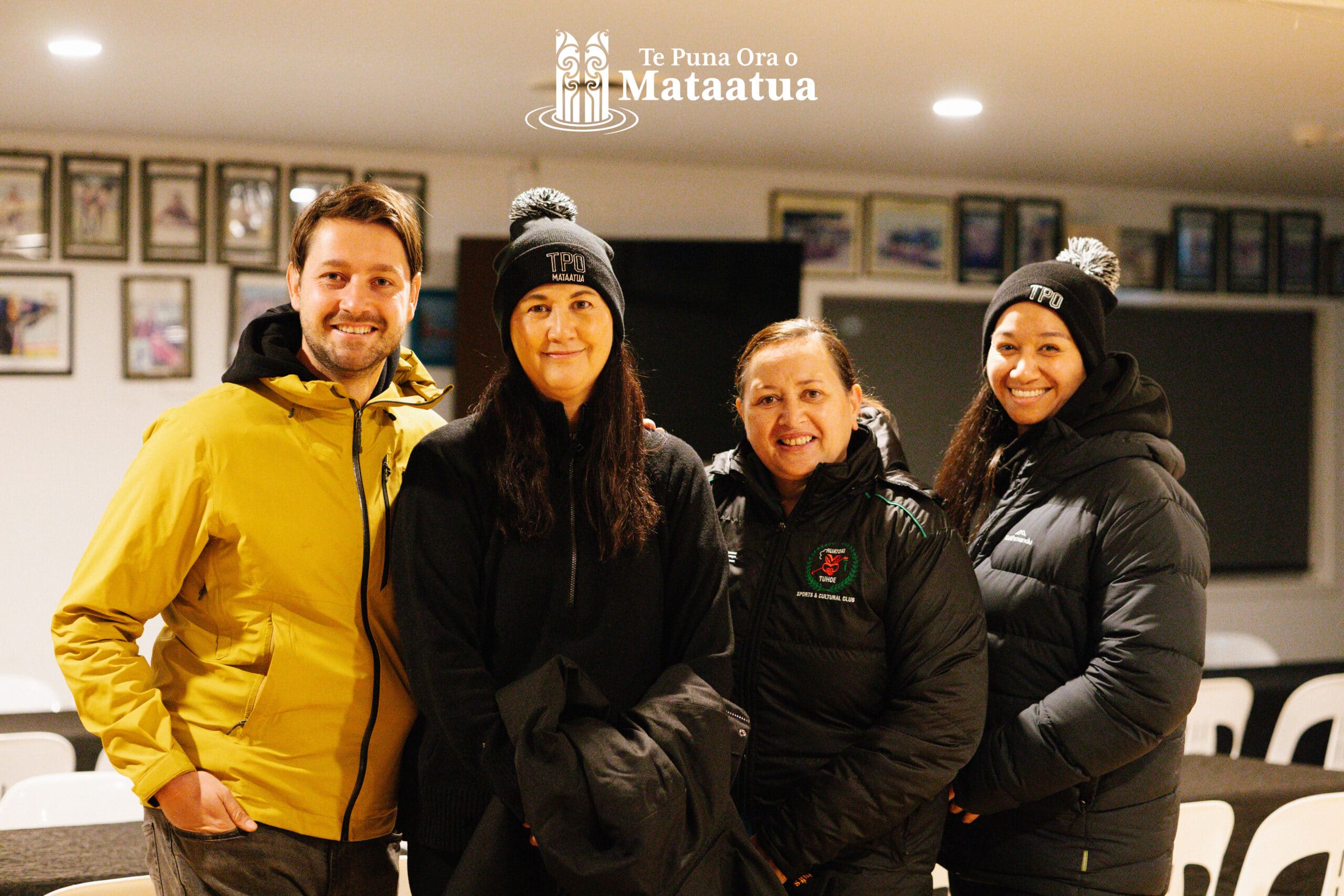 A group of people smile inside of a building in the early morning. They are all dressed in black except for one man on the far left who wears a yellow raincoat