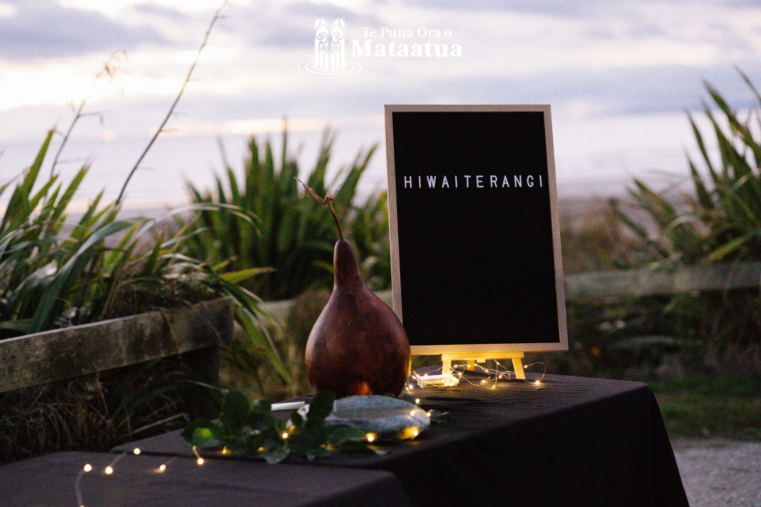 A view of a decorative table looking out over an ocean There is a gourd, a letterboard sign that says Hiwaiterangi and fairy lights on the table