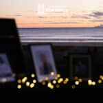 A view of a morning sunrise over the ocean. In the distance an island lightly steams and there are clouds in the pastel sky. In the foreground blurred framed photos are light by the flow of fairy lights