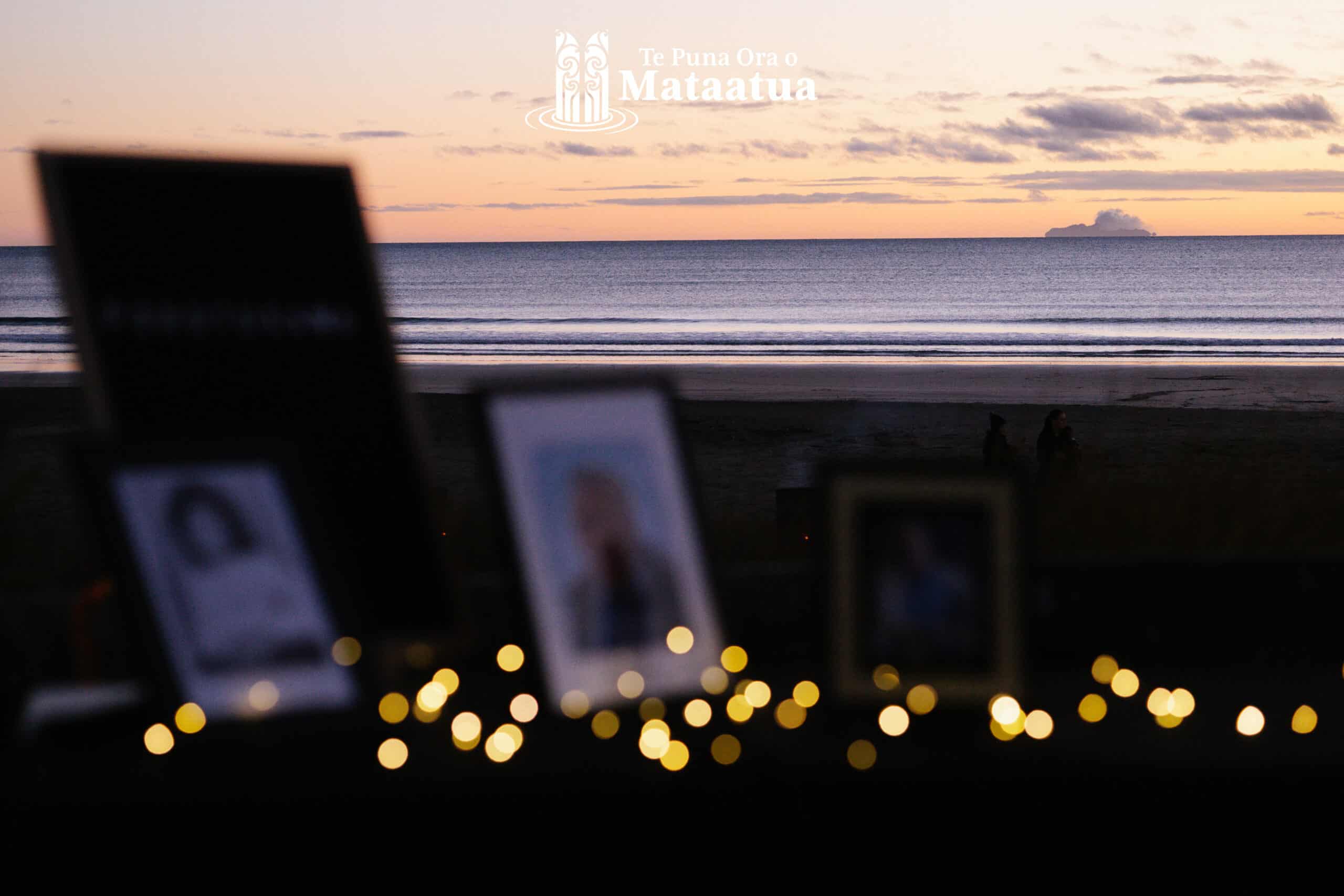 A view of a morning sunrise over the ocean. In the distance an island lightly steams and there are clouds in the pastel sky. In the foreground blurred framed photos are light by the flow of fairy lights