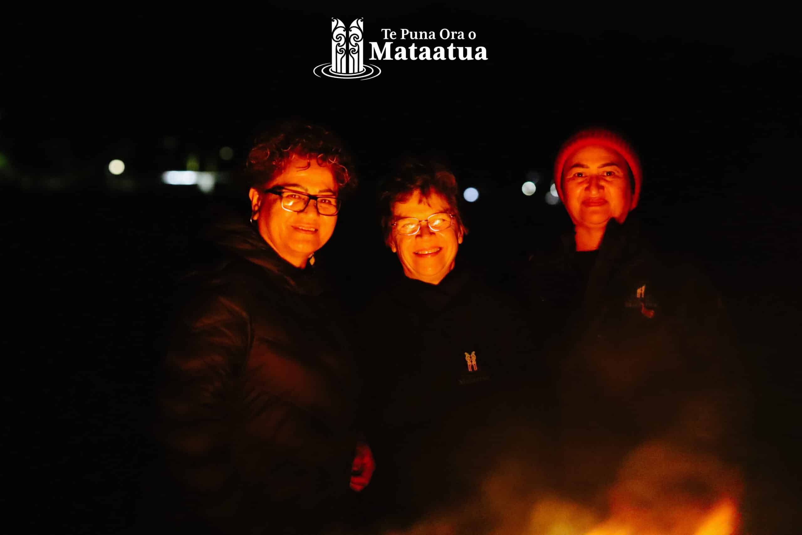 Three women huddled in front of a bonfire