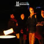 A group of people stand in front of a bonfire. Their faces are warmly lit by the fire