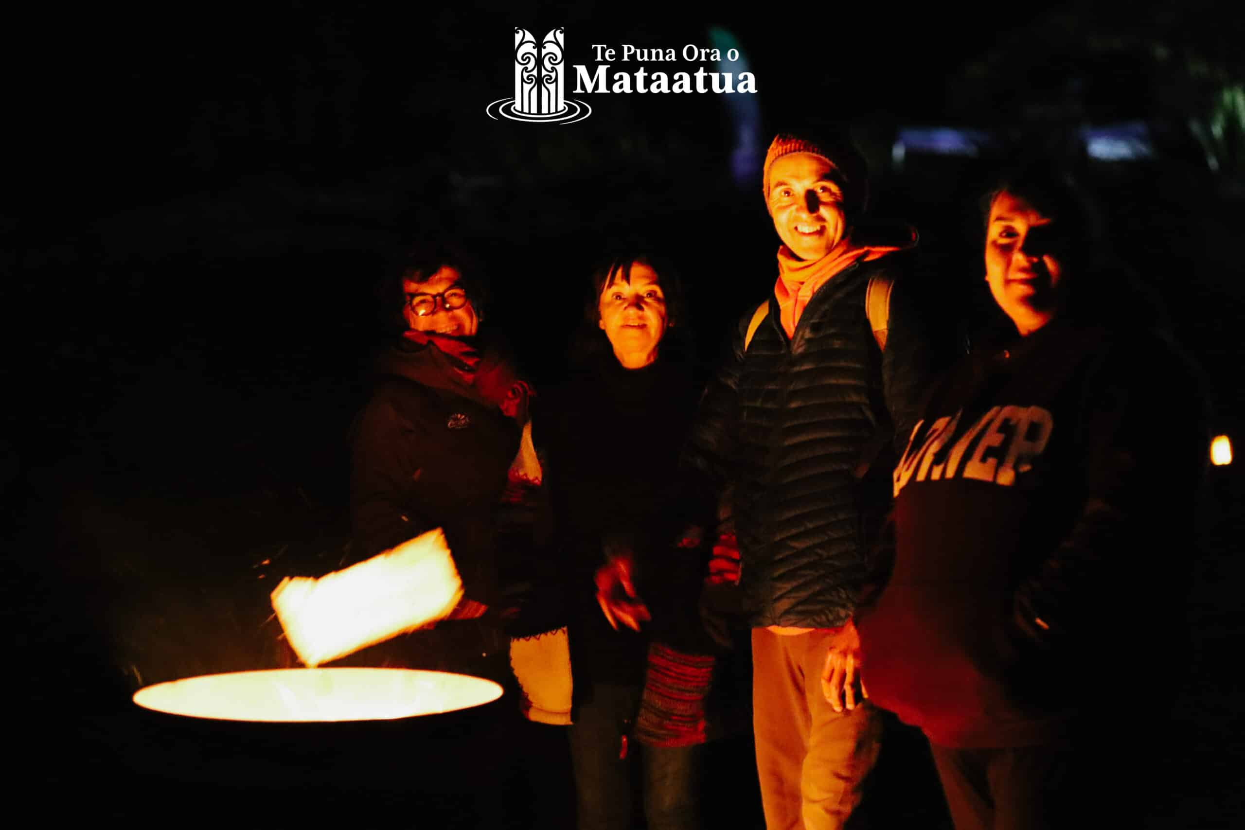 A group of people stand in front of a bonfire. Their faces are warmly lit by the fire