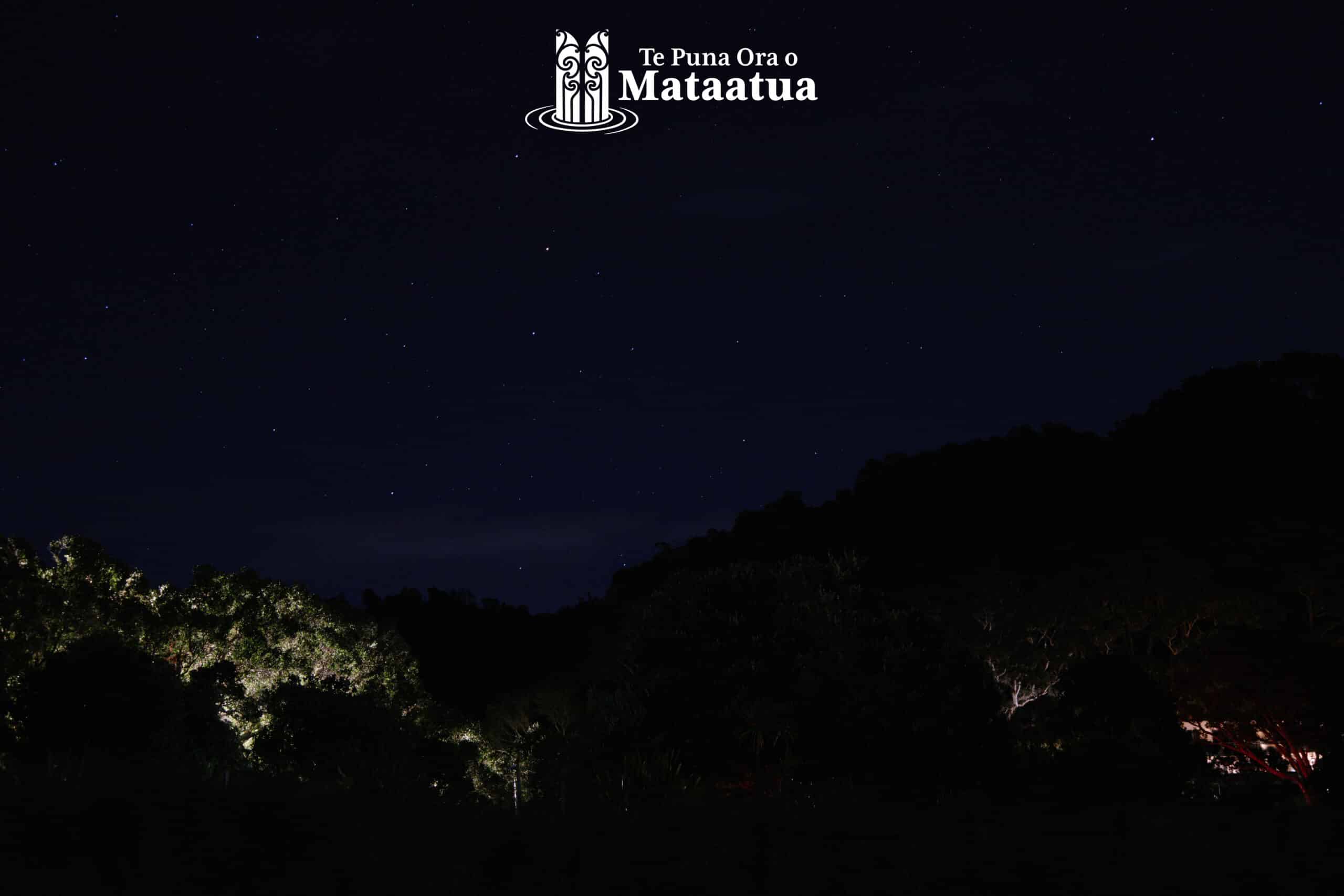 A beach scene in the early morning. At the top of the image the sky is dark blue, and there are stars peeking through thick fluffy clouds. At the bottom of the image, you can see a ridgeline of native bush, some trees are illuminated by street lights