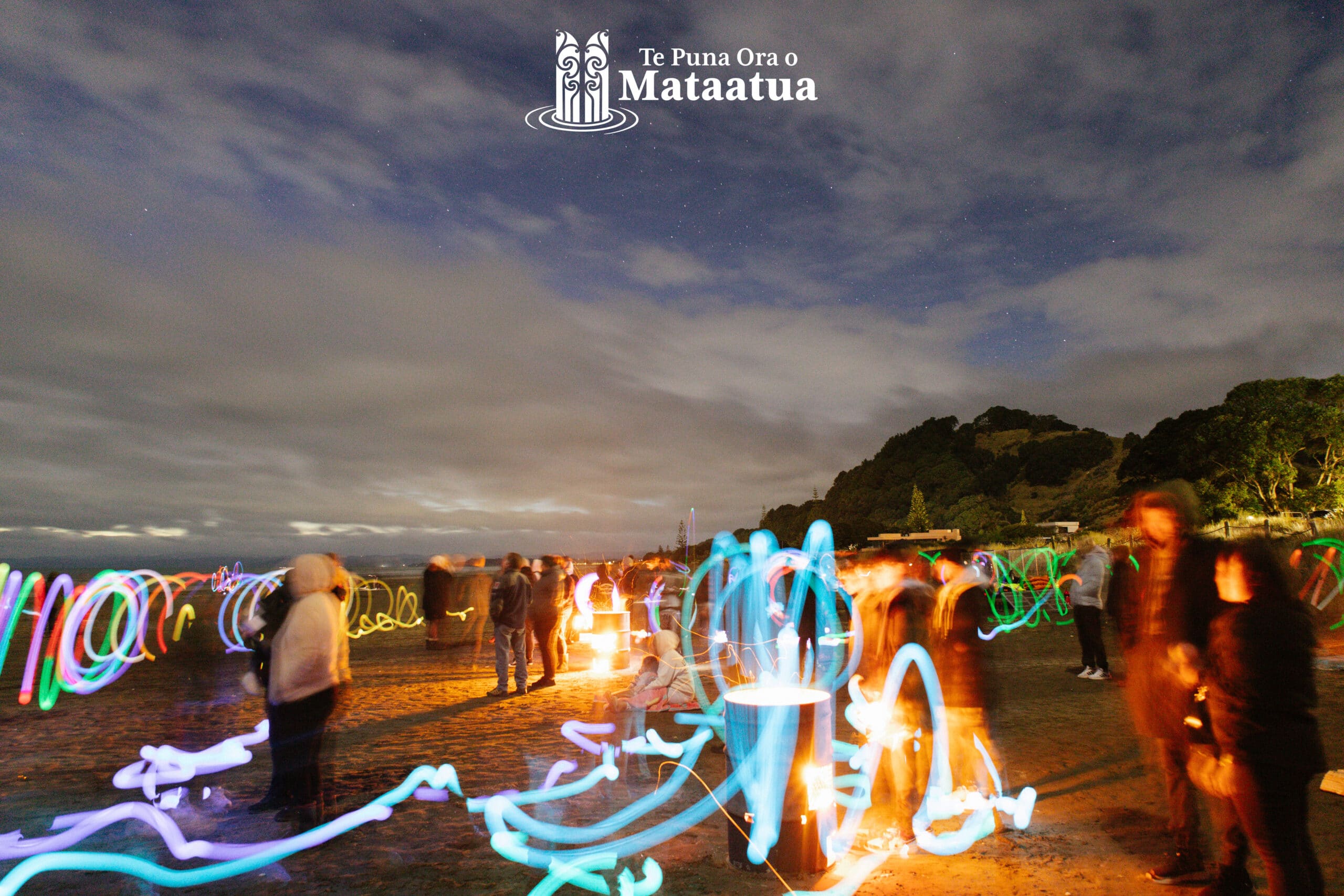 A beach scene in the early morning. At the top of the image the sky is dark blue, and there are stars peeking through thick fluffy clouds. At the centre of the image, you can see people standing by fires The image is full of many rainbow patterns of light from a long exposure of moving LED sticks