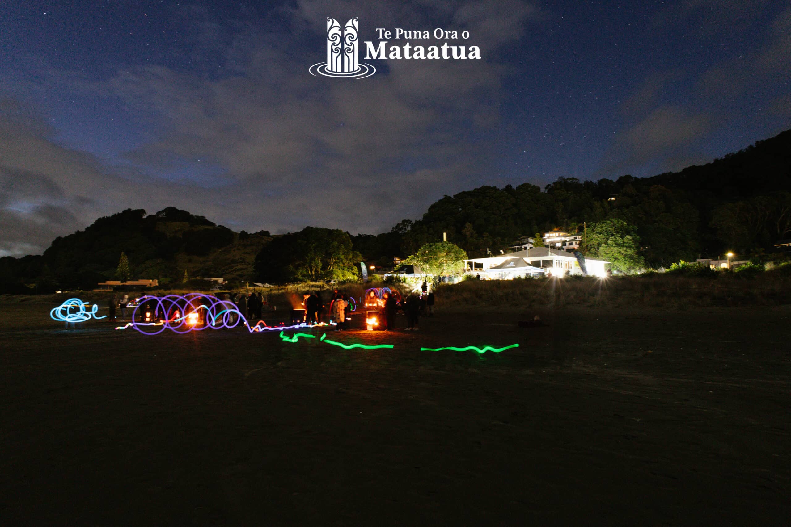 A beach scene in the early morning. At the top of the image the sky is dark blue, and there are stars peeking through thick fluffy clouds. At the centre of the image, you can see a ridgeline of native bush, and some buildings. The buildings are illuminated by big lights At the bottom of the image there is sand and a rainbow patterns of light from a long exposure of moving LED sticks