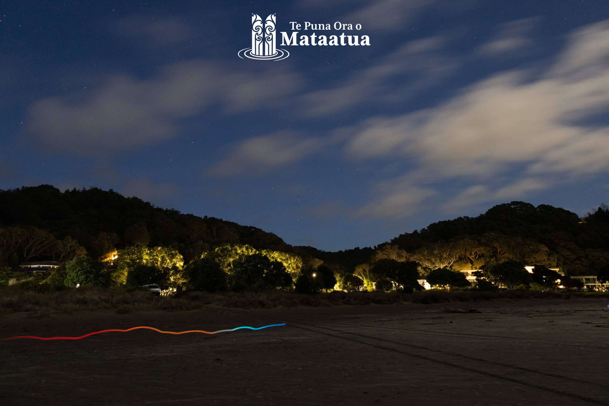 A beach scene in the early morning. At the top of the image the sky is dark blue, and there are stars peeking through thick fluffy clouds. At the centre of the image, you can see a ridgeline of native bush, some trees are illuminated by street lights At the bottom of the image there is sand and a rainbow stripe of light from a long exposure of a moving LED stick