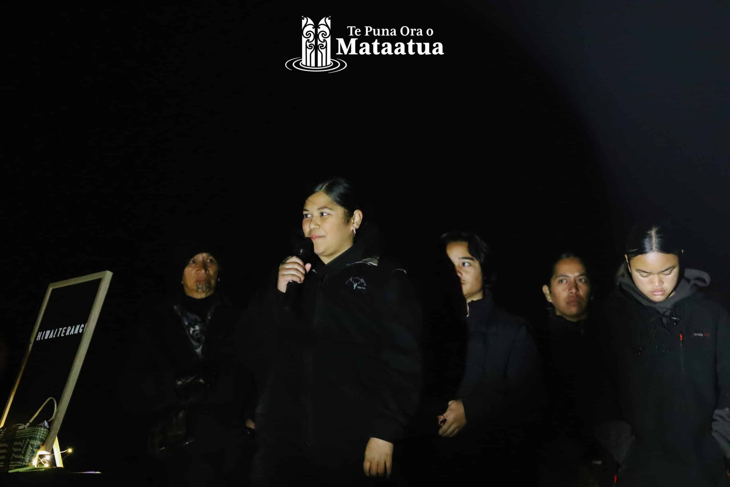 A group of children watch on as a small girl dressed in black gives a karakia/prayer