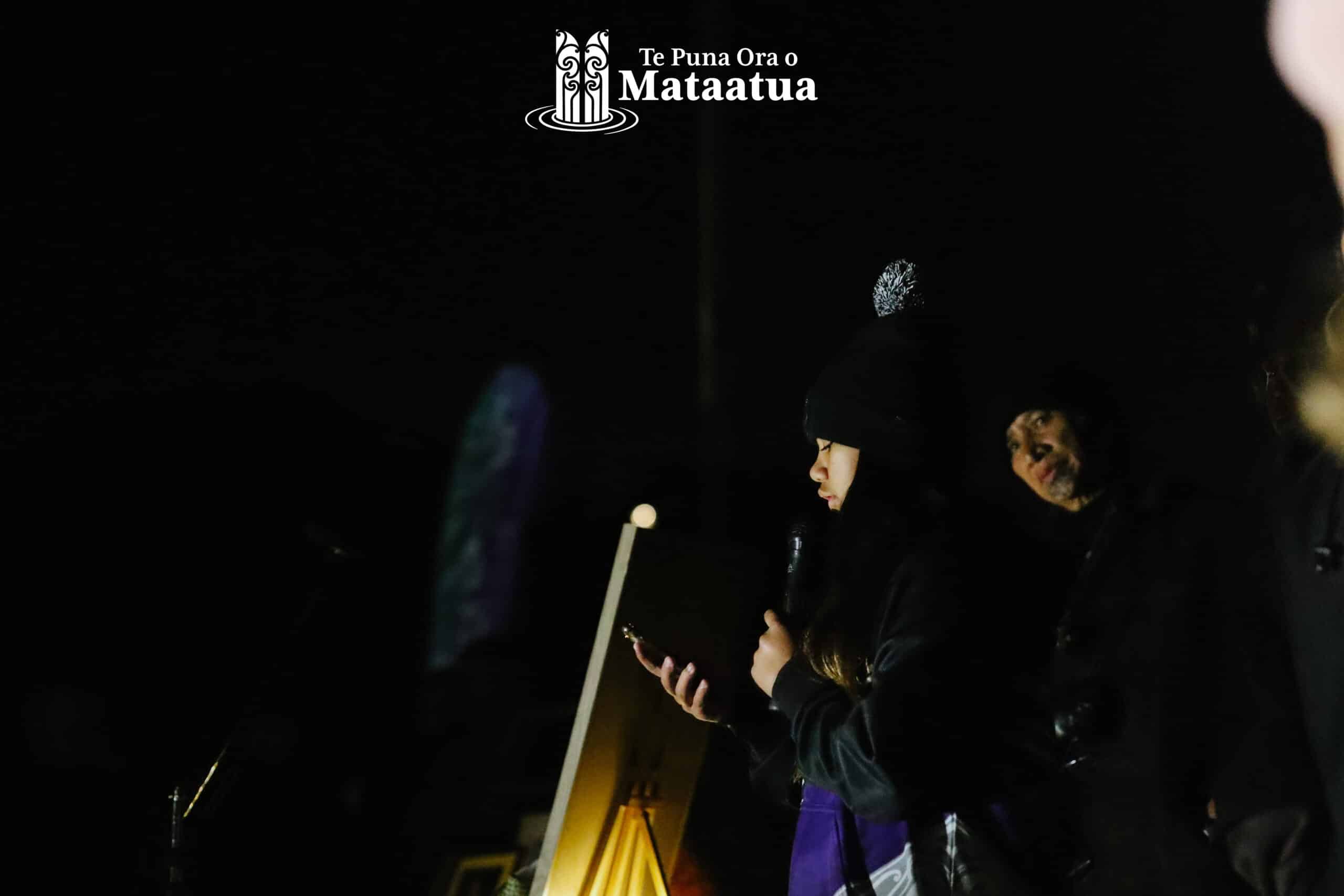 A group of children watch on as a small girl dressed in black and purple gives a karakia/prayer