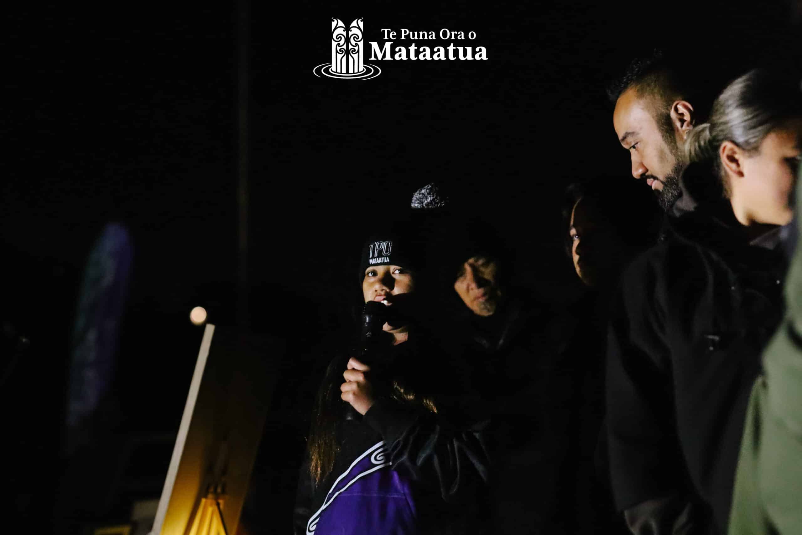 A group of children watch on as a small girl dressed in black and purple gives a karakia/prayer