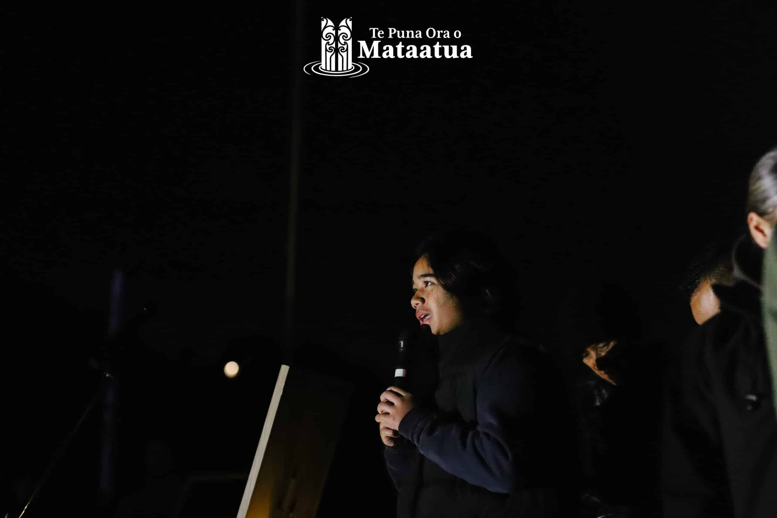 A group of children watch on as a small girl dressed in black gives a karakia/prayer