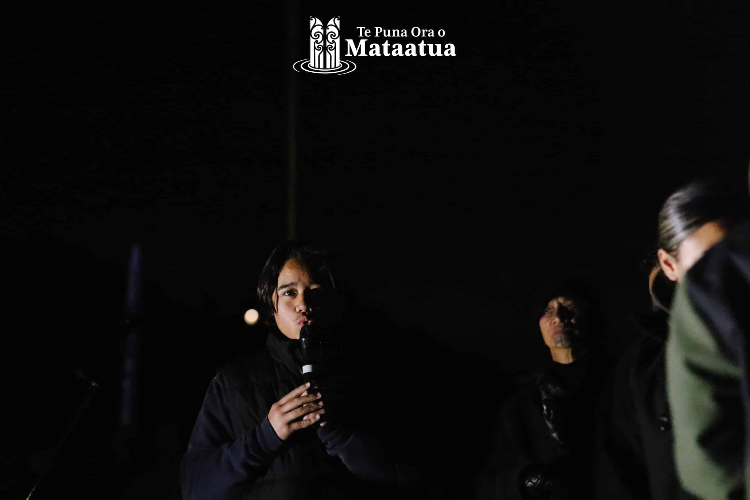 A group of children watch on as a small boy dressed in black gives a karakia/prayer