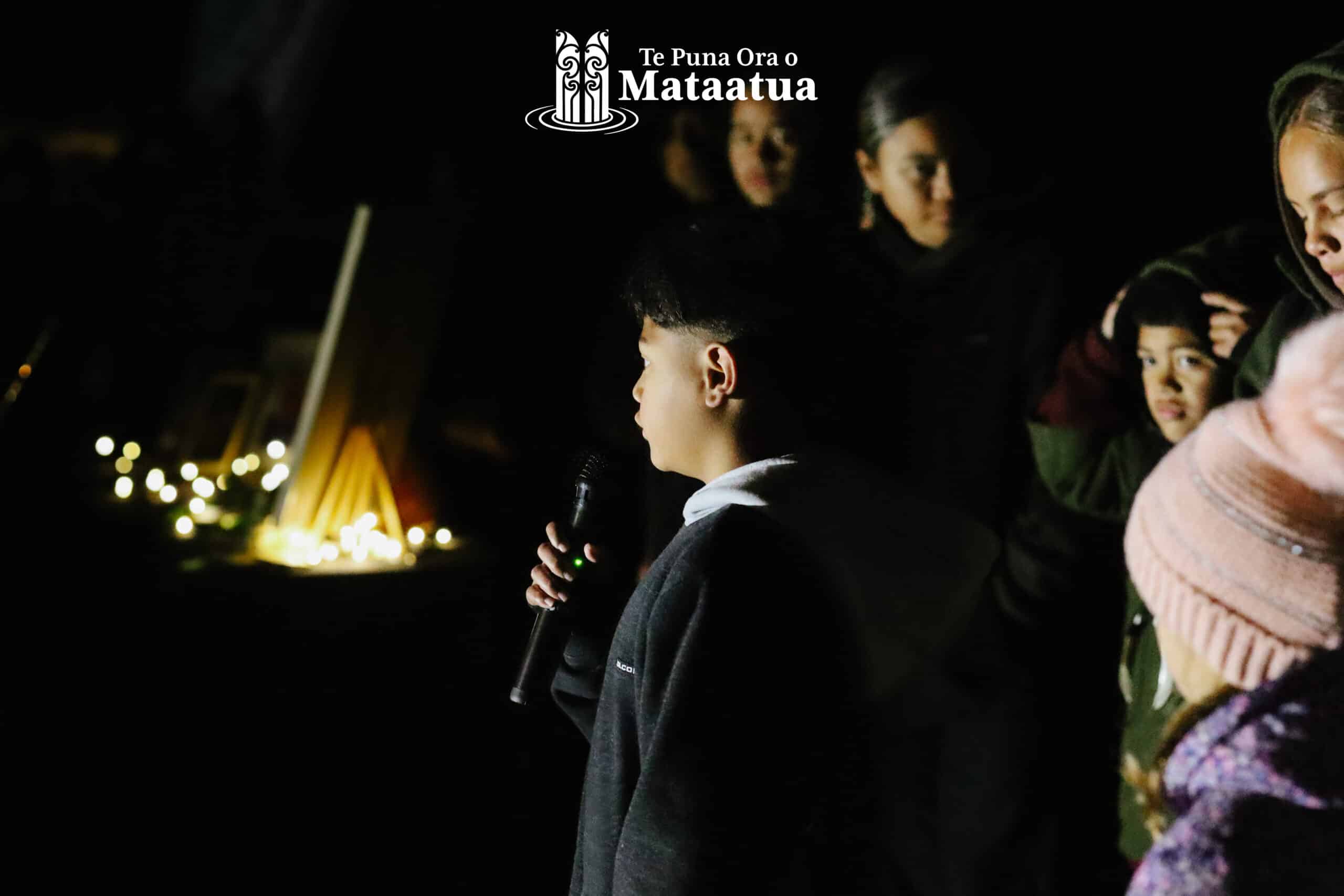 A group of children watch on as a small boy dressed in black gives a karakia/prayer