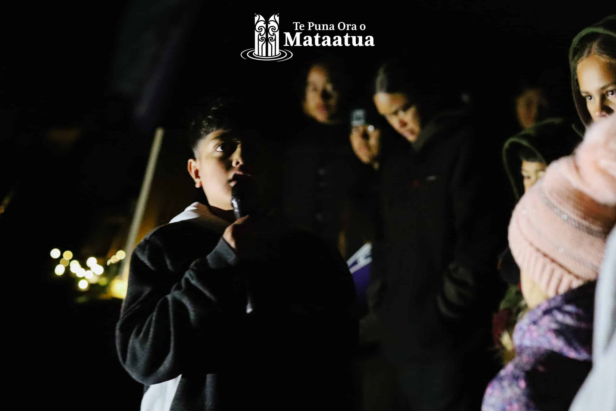 A group of children watch on as a small boy dressed in black gives a karakia/prayer