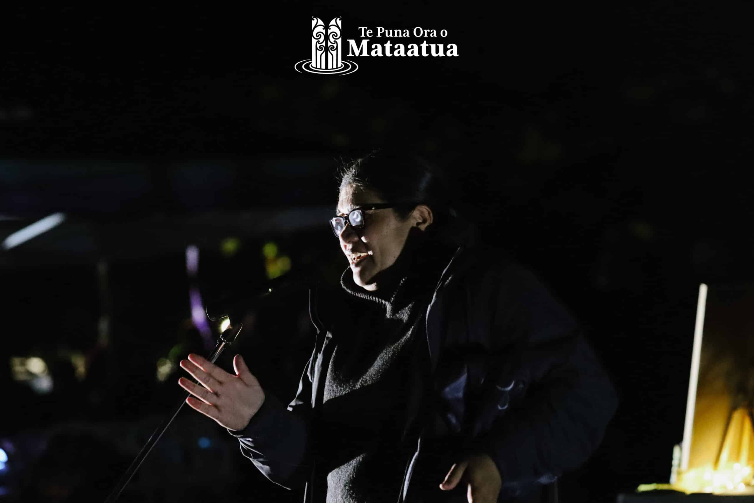 A woman gives a presentation on Matariki in the darkness of early dawn. Her face is lightly illumated by a distant flood light