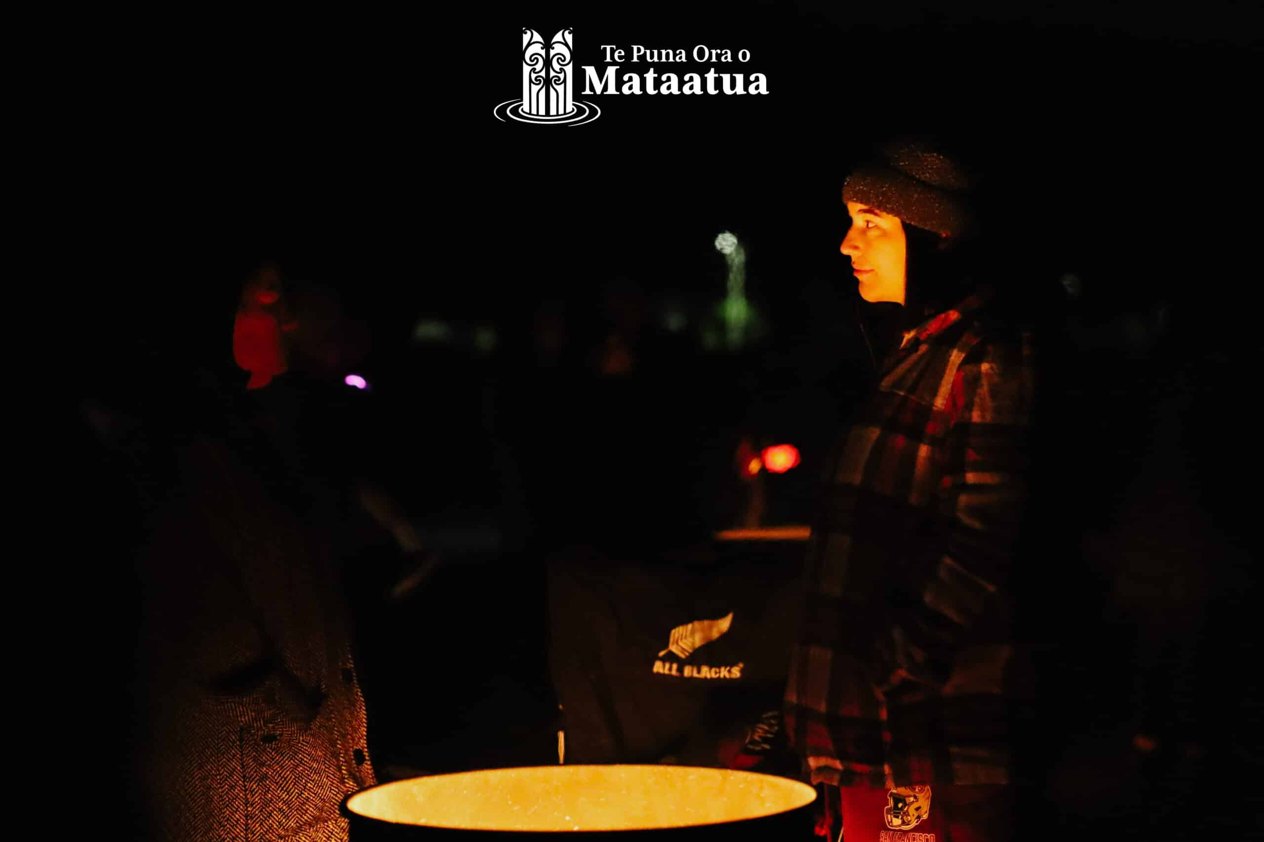 A woman ponders over a bonfire at dawn. Her face is lightly lit by the flow of the flames beneath her