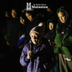 A group of children watch as a young girl dressed in purple gives a karakia