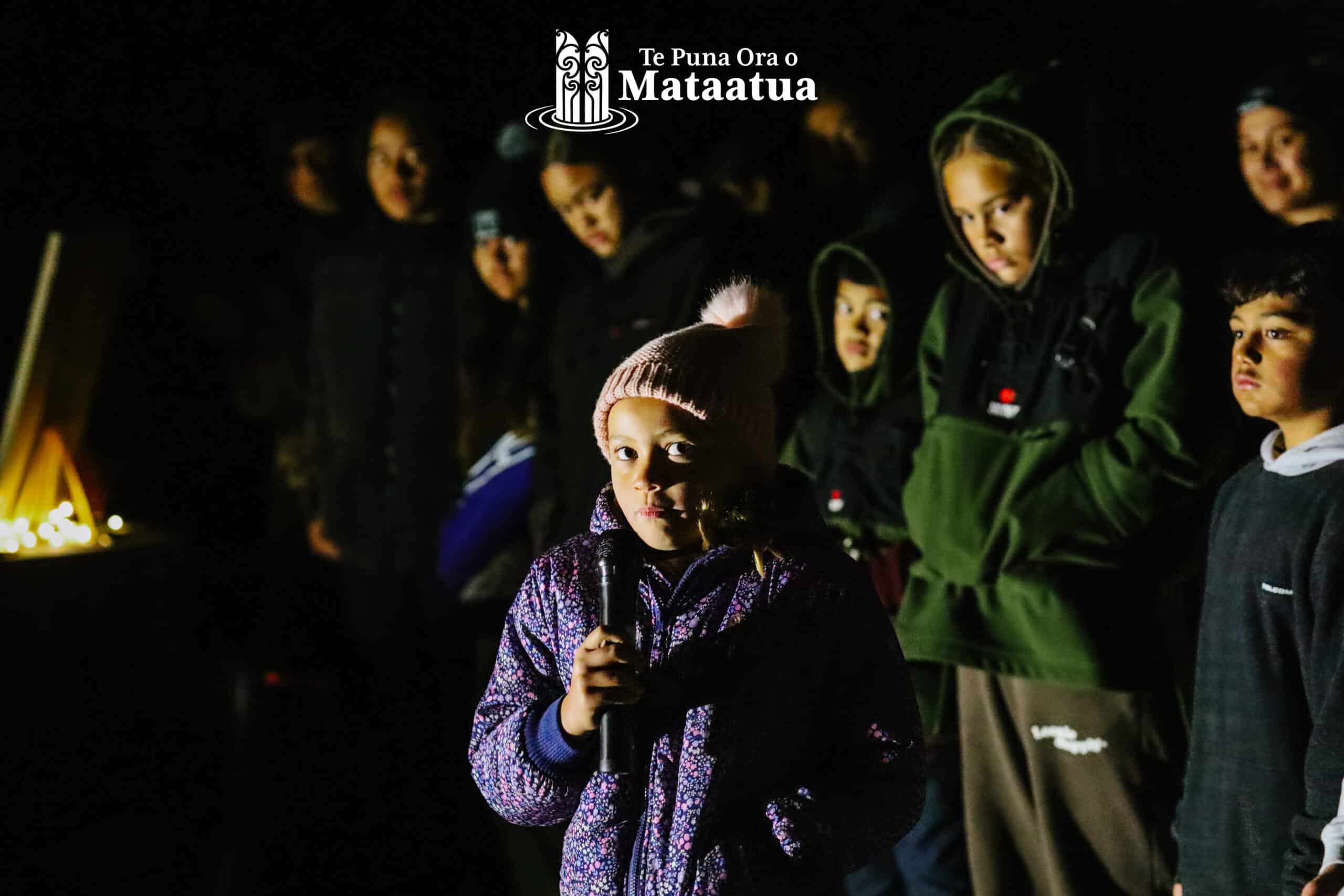 A group of children watch as a young girl dressed in purple gives a karakia