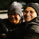 Two women in winter clothing smile, their faces lit by a warm light