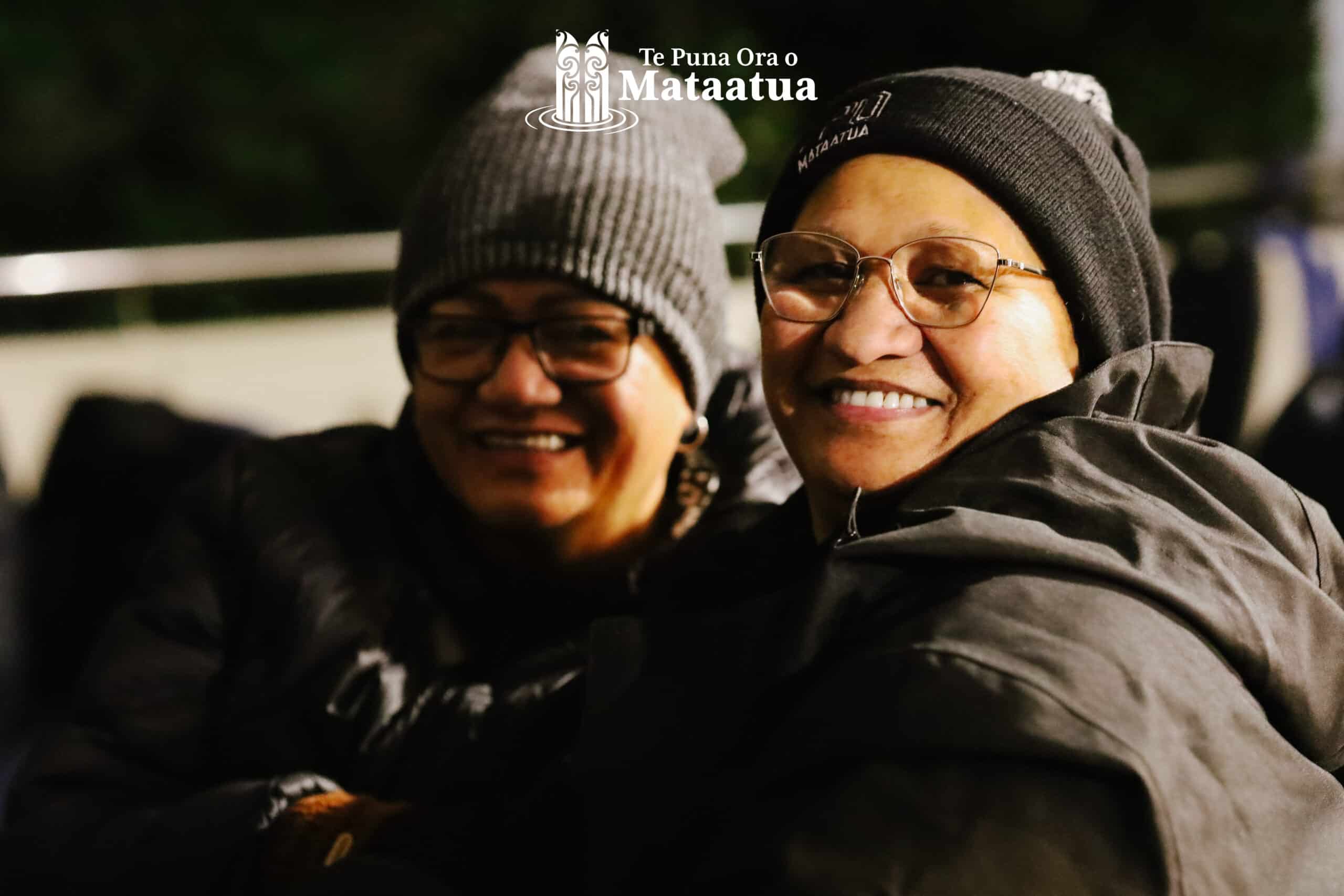 Two women in winter clothing smile, their faces lit by a warm light