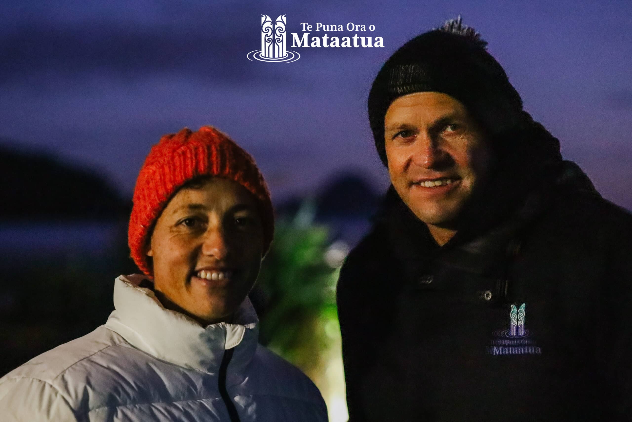 A man and a woman smile. The image is darkly lit and taken in the early evening