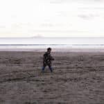 A young boy walks alone on a beach against a pastel sunrise. In the background a volcanic island lightly steams on the horizon