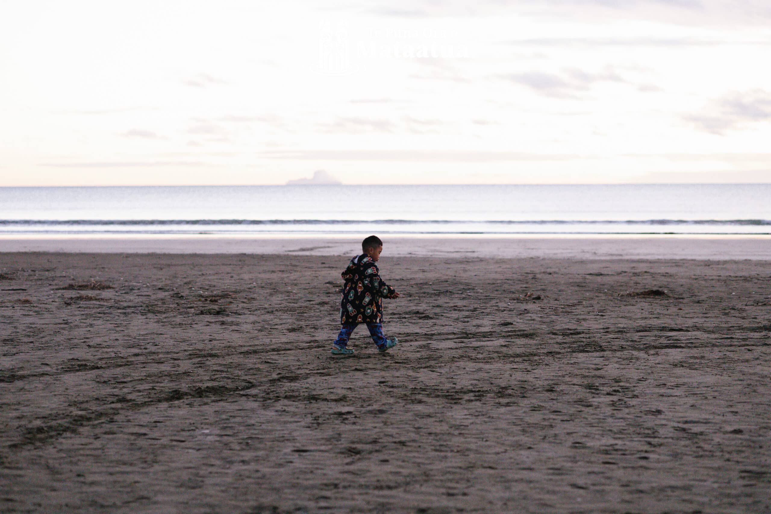 A young boy walks alone on a beach against a pastel sunrise. In the background a volcanic island lightly steams on the horizon