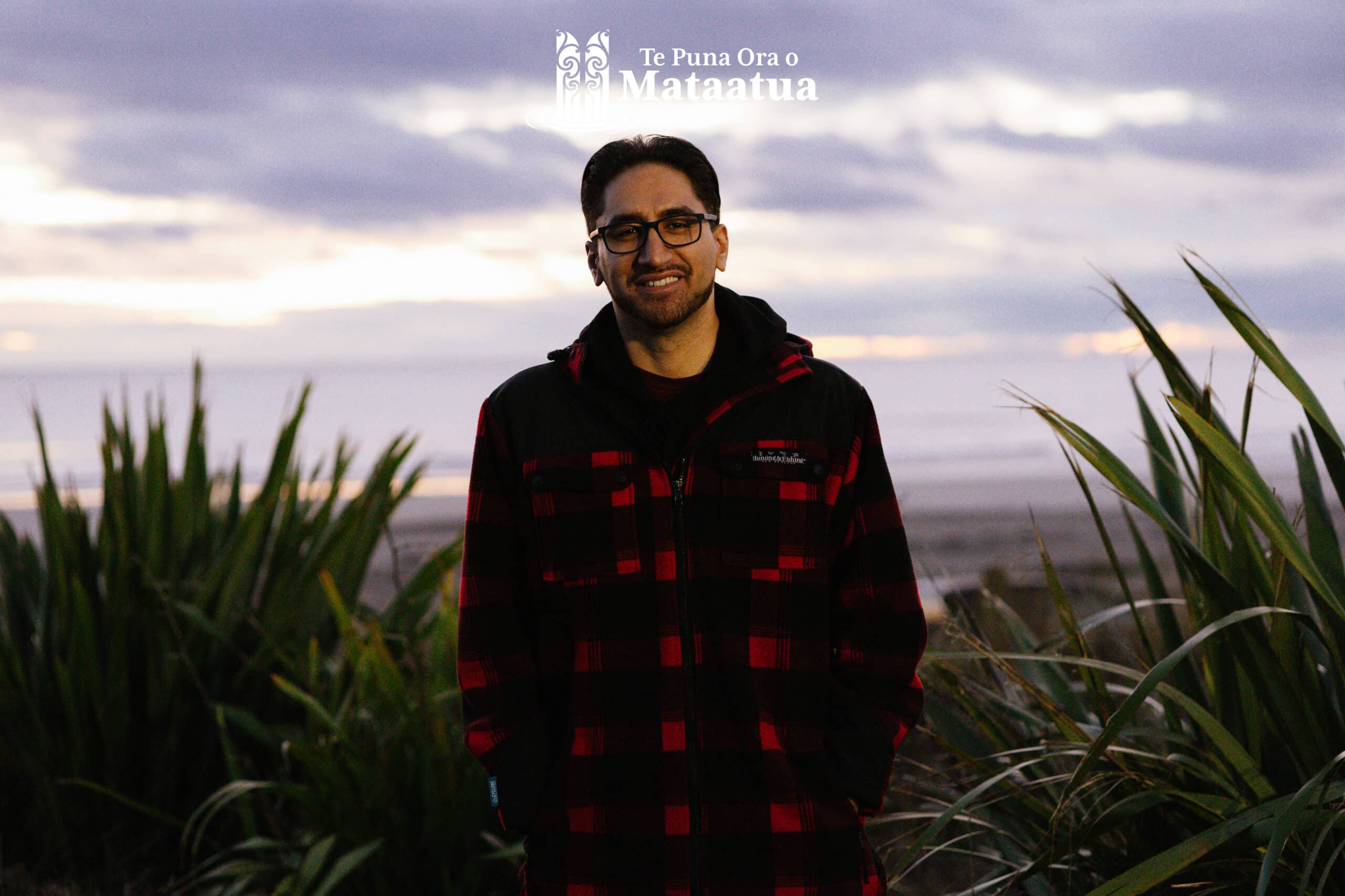 A man standing amongst green flax, against a cloudy pastel sunrise.