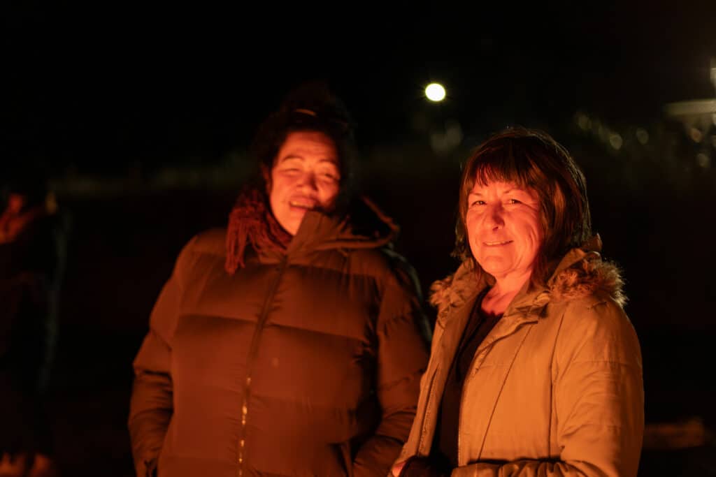 Two woman smiling their faces are warmly lit by a bonfire