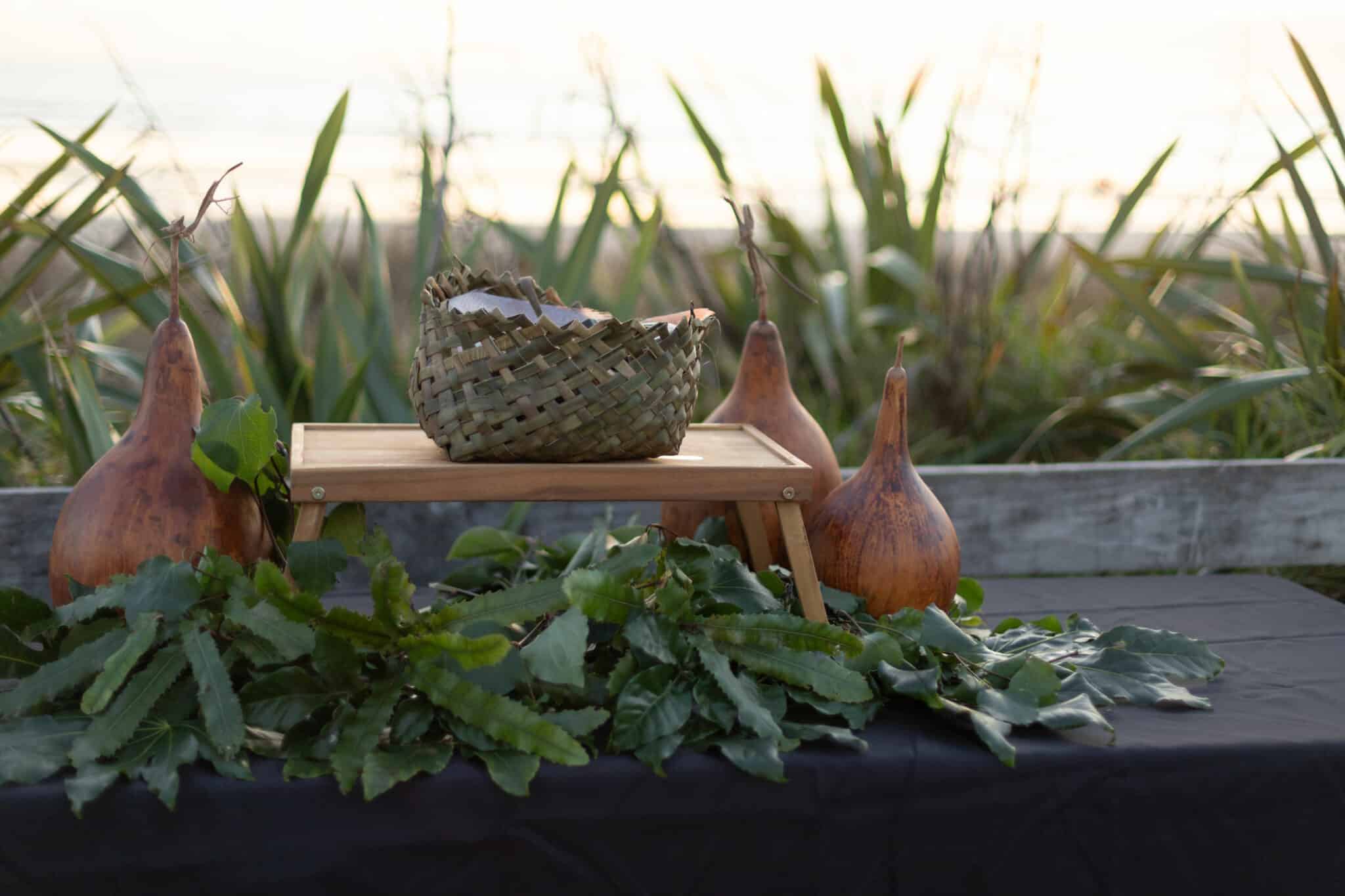 A small flax basket on a stand, decorated by kawakawa leaves and surrounded by hue