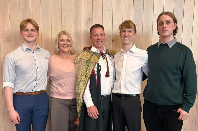 A family stands in an embrace. The man in the middle is wearing a korowai (Māori cloak)